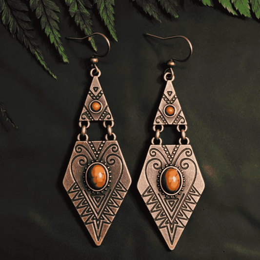 A pair of oxidised tribal stone drop earrings with bohemian dangle design, featuring brown stones and intricate patterns, laid on a dark surface with green foliage.