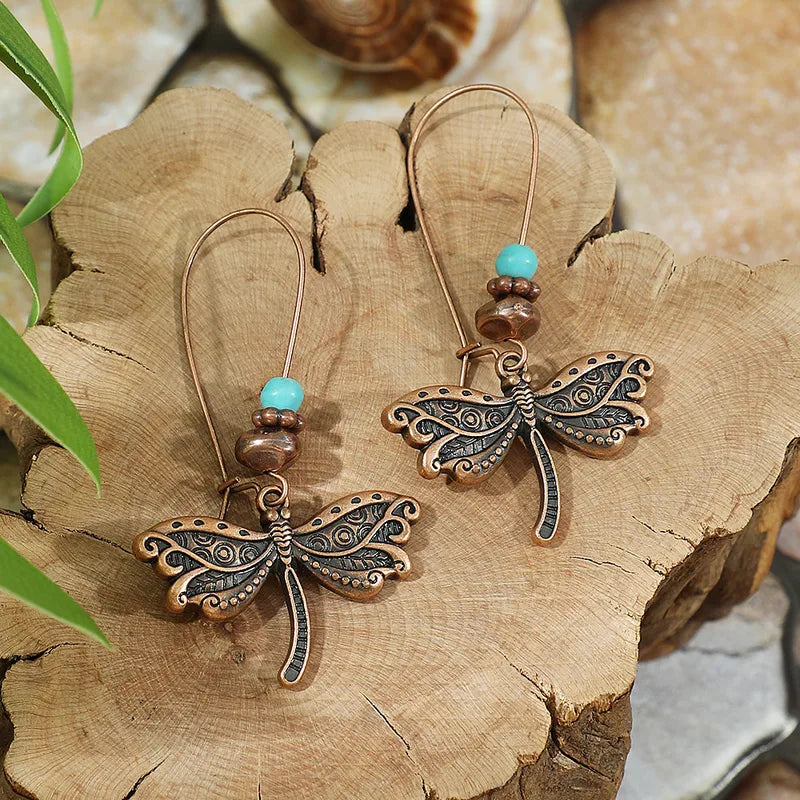 Pair of oxidized bronze dragonfly dangle earrings with turquoise and copper beads on a natural wooden surface.