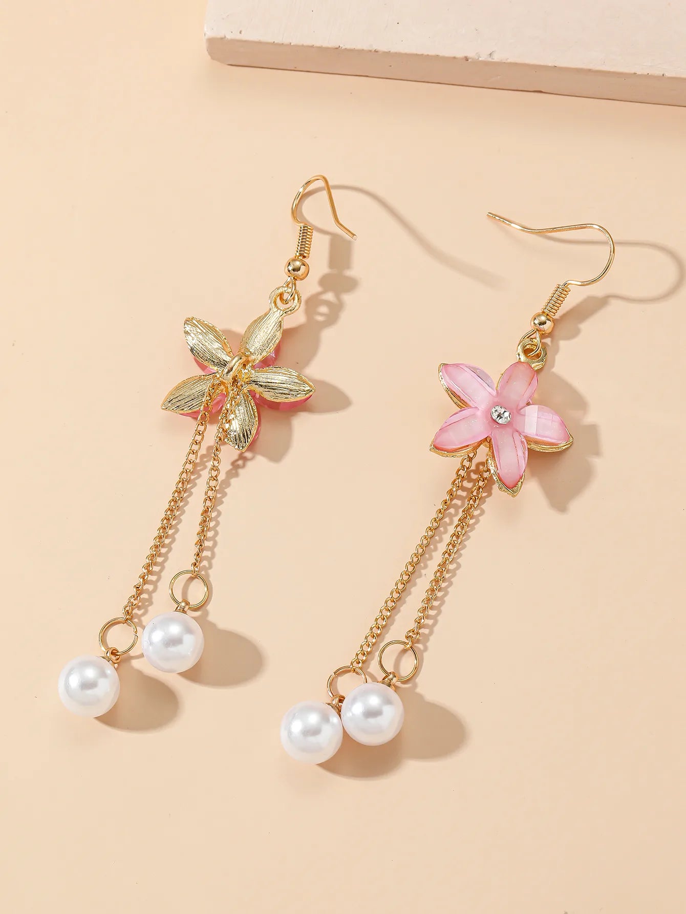 A pair of pink floral drop earrings with pearl chains lying on a beige surface, showing the front and back of one earring.