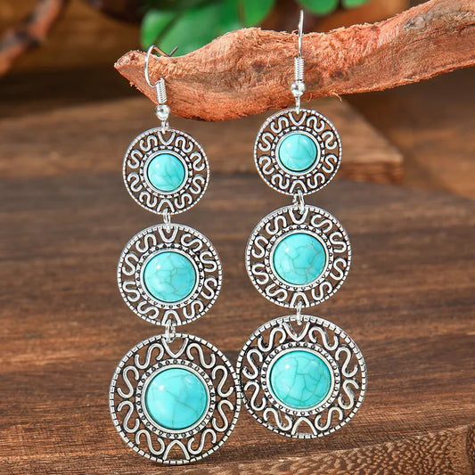 Pair of long, tiered turquoise chandelier earrings with oxidised silver-tone metal and scroll patterns, hanging from a natura