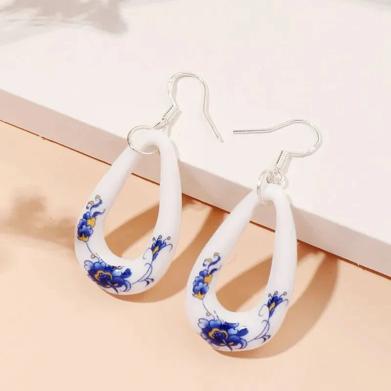 A pair of white ceramic drop earrings with blue and yellow floral patterns, featuring silver-toned hooks, displayed on a light background.