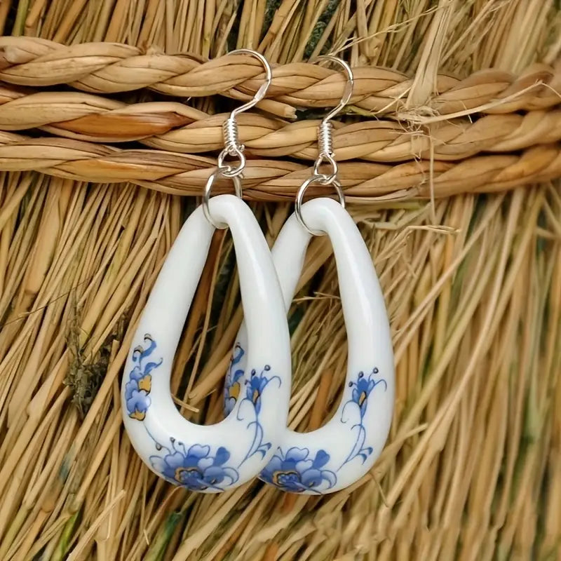 A pair of white ceramic drop earrings with blue and yellow floral patterns, featuring silver-toned hooks, hanging against a rustic straw background.